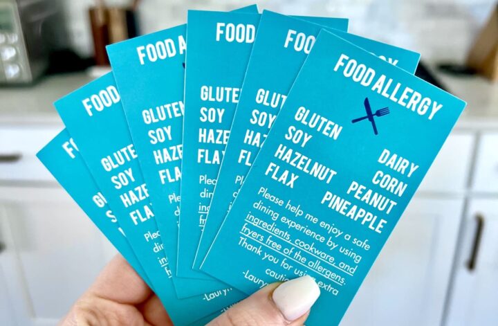 Holding food allergy cards for gluten, dairy, soy, corn, hazelnut, peanut, pineapple, and flax allergies.